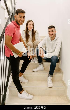 Cheerful young multiethnic male and female friends sitting on staircase of university building with laptop Stock Photo
