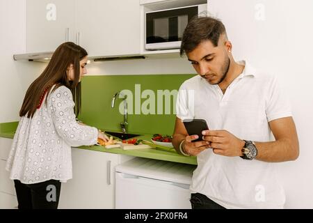 Concentrated young ethnic guy messaging on smartphone standing in kitchen near girlfriend preparing fresh healthy salad Stock Photo