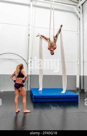 Full body fit female in sportswear looking at muscular male partner performing upside down exercises on aerial silks while training together in modern Stock Photo