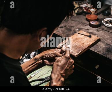 From above cropped unrecognizable concentrated ethnic elderly male jeweler in casual clothes and eyeglasses sharpening instruments while working at wo Stock Photo