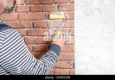 Crop anonymous bearded middles aged male master in casual clothes applying glue on brick wall with roller during renovation works in apartment Stock Photo