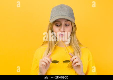 Perplexed Woman in yellow sweater and cap holding stylish sunglasses while standing on yellow background Stock Photo