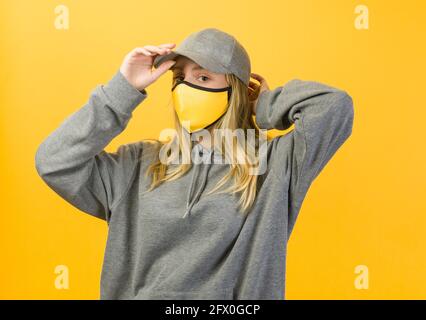 Unemotional young female in loose hoodie and yellow face mask putting on cap and looking at camera against yellow wall Stock Photo