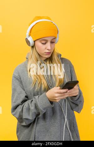 Focused young female in casual wear listening to music in headphones and browsing mobile phone against yellow background Stock Photo