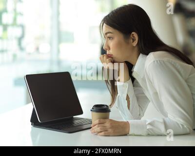 Side view of young female office worker hand to cheek ​and holding paper cup while thinking about her work Stock Photo