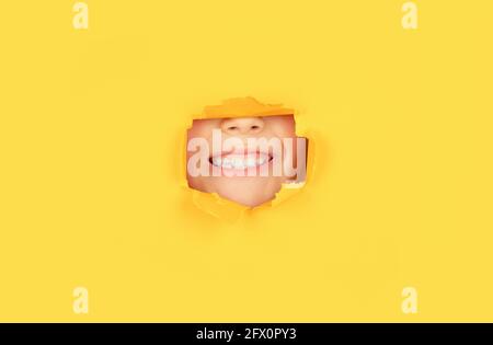 Positive child with toothy pleasant smile on face, keeps through torn hole in yellow paper. Breaking paper background. Emotions concept. Kid with Stock Photo