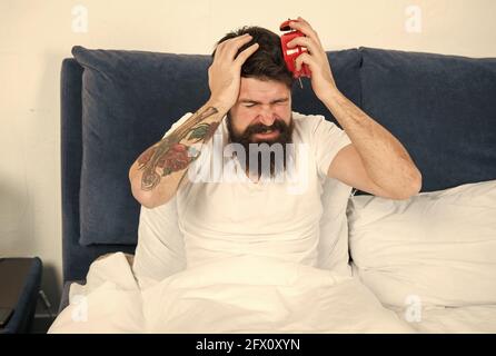My clock is running late. Late riser suffer headache. Stressed hipster hold alarm clock in bed. Bearded man wake up late in morning. Keeping late Stock Photo
