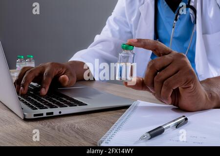 Mid section of african american male doctor using laptop and holding covid-19 vaccine bottle Stock Photo