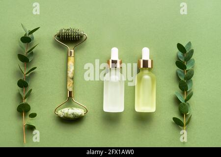 Green facial jade massage roller, glass dropper bottles for medical and cosmetic use, and eucalyptus on a green background. SPA concept. Stock Photo