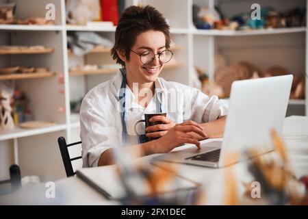 Happy woman in glasses using laptop touchpad in pottery shop Stock Photo