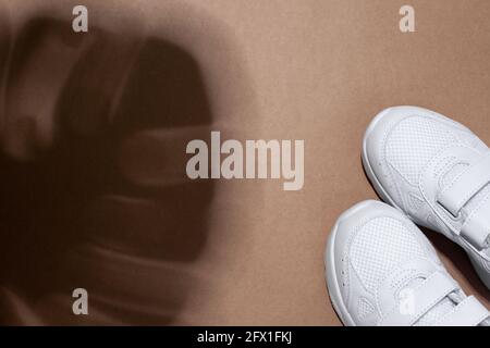 top view two white women's sneakers in the corner with shadows from a monstera leaf isolated on a beige craft paper background Stock Photo