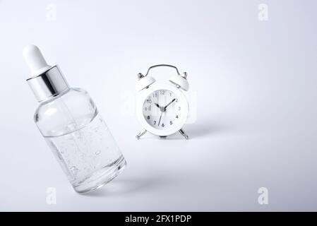 Bottle of face serum levitates on a white background next to the retro alarm clock. The concept of leaving youth. Maintain skin firmness. Stock Photo