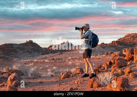 Photographer taking photo in rocks of Namib Desert, Namibia, Africa. Red mountains and sunset sky in background. Landscape photography