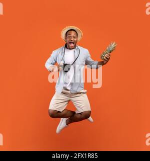 Full length of African American man in summer clothes holding camera and pineapple, jumping on orange background Stock Photo