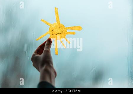 A child's hand holds the sun, cutted out of felt, against the window. Outside the window, rain and bad weather. Close-up. The concept of depression an Stock Photo