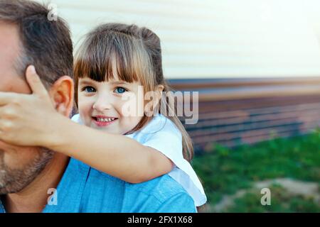 Happy little girl hugs dad on the street. Child plays with parent in hide and seek. The baby is smiling outdoors. Stock Photo