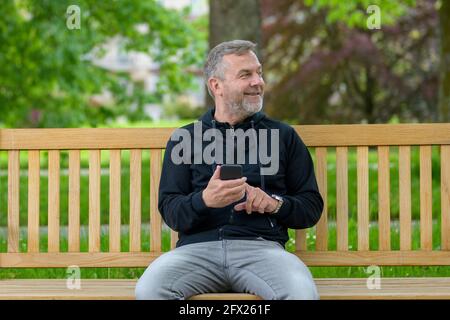 Happy relaxed middle-aged man using his mobile phone in a park texting a message while looking to the side Stock Photo