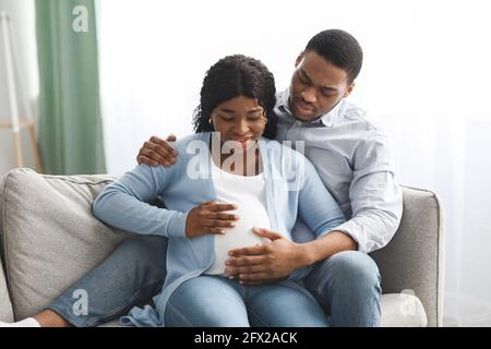 Worried expecting family sitting on couch, touching big belly Stock Photo