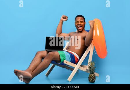 Black man showing laptop with empty screen while relaxing in lounge chair, gesturing YES on blue background, mockup Stock Photo
