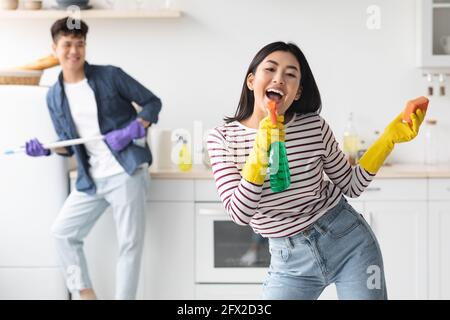 Joyful asian lady using spray as microphone, singing while cleaning Stock Photo