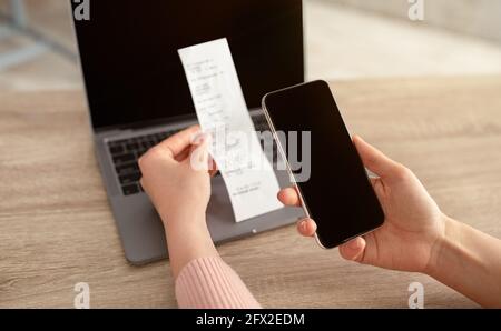 Female hands holding smartphone with empty screen and bill or slip, sitting at workplace with laptop on desk Stock Photo