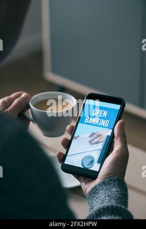 closeup of a young caucasian man, sitting at a coffee table, having a cup of coffee, watching at his smartphone, that reads the text open banking Stock Photo