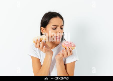 Portrait young beautiful woman craving to eat donuts licking her lips on white studio background. Stock Photo