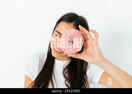 Portrait of happy young woman looking through hole in pink glazed donut on white studio background. Stock Photo