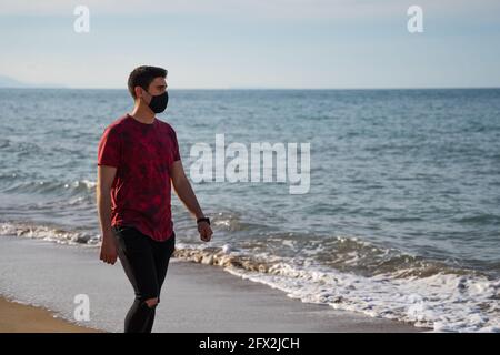 A man walking along the seashore barefoot. Caucasian man in red T-shirt and black trousers. On a sunny day on the sandy beach. Wearing a watch and mas Stock Photo