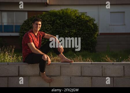 Man sitting on a wall smiling. He is barefoot and wearing a watch. He is wearing a red short-sleeved T-shirt and black trousers. Stock Photo