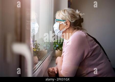 Senior Woman in Protective Face Mask Looking Through Window at Home Stock Photo