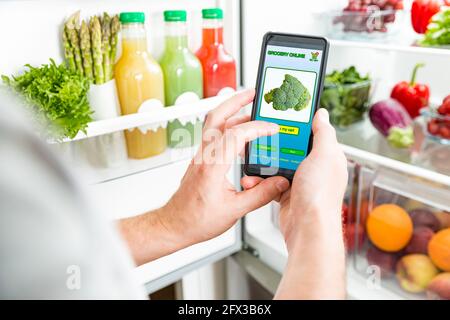 Man using smart phone, buying food. Close-up hand with phone. Grocery shopping online. Mobile app of online food store. Fresh vegetables in fridge Stock Photo