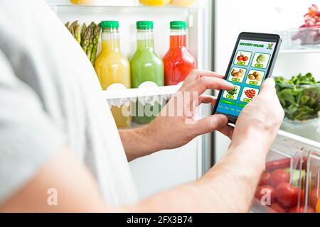 Man using smart phone, buying food. Close-up hand with phone. Grocery shopping online. Mobile app of online food store. Fresh vegetables in fridge Stock Photo
