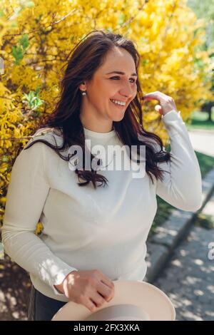 Portrait of smiling middle-aged woman walking in blooming spring garden. Happy stylish lady holding hat outdoors surrounded with flowers Stock Photo