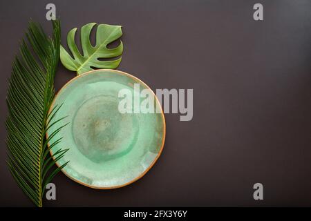Empty green plate on black background. Luxury dinner plate served with asian kinfolk grunge style tropical palm leaves monstera plant. Mock up empty Stock Photo