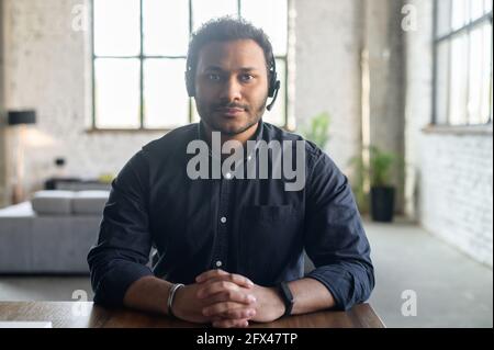 Portrait of serious indian guy wearing wireless headset looks at the camera, video connect with multiracial colleague, video chat with hindu guy in headphones, webcam view, video call concept Stock Photo