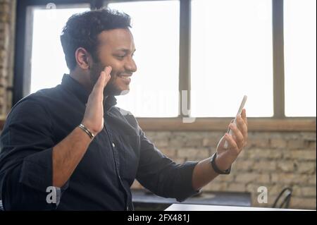 Indian guy using mobile app for video connection, holding smartphone and waving to online interlocutor, happy hindu guy enjoying chatting via video call, staying in touch with family on the distance Stock Photo