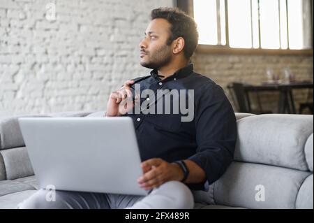 Contemplative mixed-race hindu guy sits on the sofa with a laptop and waiting for inspiration, looks away, figured out how to solve an important problem or project issues Stock Photo