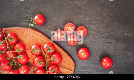 Fresh red cherry tomatoes on wooden cutting board, some sliced to half over black slate like board near Stock Photo