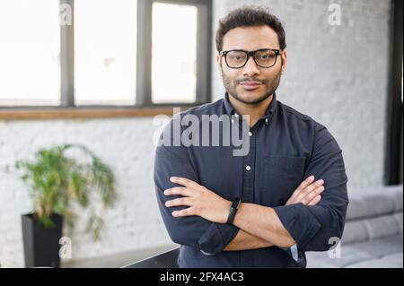 Ambitious hindu man in smart casual shirt stands with arms crossed in contemporary office space and looks at camera, portrait of purposeful indian businessman, ceo indoor. Diversity of office staff Stock Photo