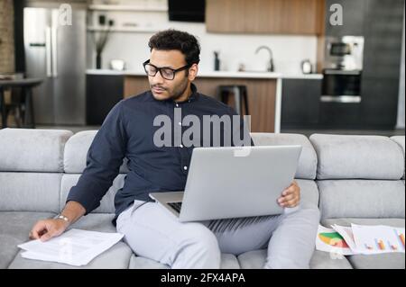 Mixed-race indian man in casual shirt sits on the couch and using laptop for remote work, focused freelance guy wearing eyeglasses doing paperwork, checking report, preparing business plan Stock Photo