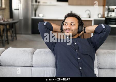 Time to relax. Indian guy in headphones sitting on the couch, holding hands behind head and listening music, hindu man with earphones enjoying audiobooks staying at home Stock Photo