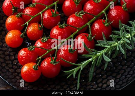 Wet ripe red cherry tomatoes on the vine and sprig of fresh rosemary on a black plate. Macro. Water drops on a small round tomatoes. Freshness, tasty Stock Photo