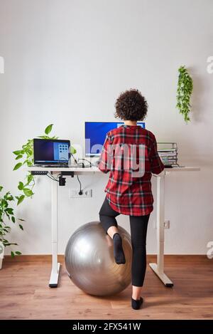 Unrecognizable woman teleworking at an adjustable standing desk with one knee resting on a fitball Stock Photo