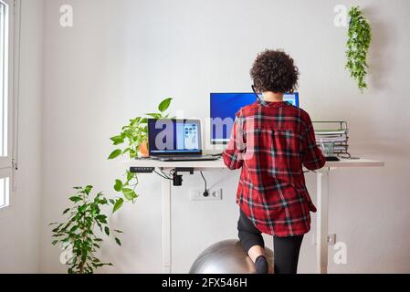 Unrecognizable woman teleworking at an adjustable standing desk with one knee resting on a fitball Stock Photo