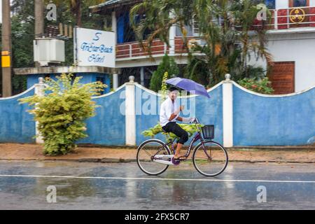 Sri Lankan male rides bicycle carrying umbrella in torrential rain (taken using 'panning' camera technique), Arugam Bay, Eastern Province, Sri Lanka Stock Photo