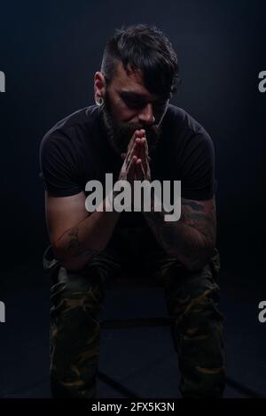 Closeup dark picture of a bearded male model in black t-shirt  sitting on a chair and praying with his tattooed hands Stock Photo