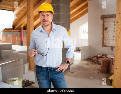 Man, builder with blue shirt and black glasses and yellow safety helmet and blue shirt standing on construction site in loft and happy and laughing Stock Photo