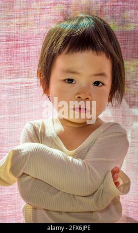 Portrait cute short hair Asian toddle girl with arms cross on the chest, standing in front of curtain, bright daylight from behind, eyes looking at ca Stock Photo