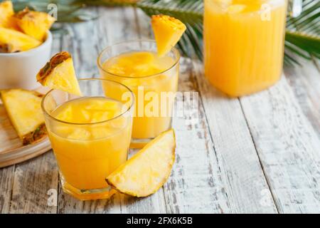 Tasty pineapple juice in two glasses with pineapple slices. Fresh natural pineapple cocktail and juice in Glass Jug on white wooden table with palm Stock Photo